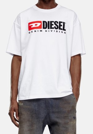 Mężczyzna w białym t-shircie Diesel Denim Division i wyblakłych szarych jeansach, stojący z jedną ręką w kieszeni na jednolitym tle.