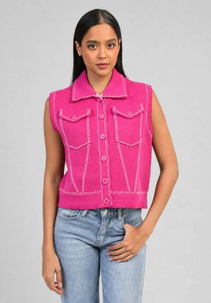 Femme portant un gilet rose sans manches boutonné avec des coutures blanches et un jean bleu, debout devant un fond gris uni.