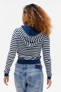 Vrouw met krullend haar, gekleed in een blauw-witte gestreepte hoodie en blauwe jeans, staande met haar gezicht naar weg tegen een effen witte achtergrond.