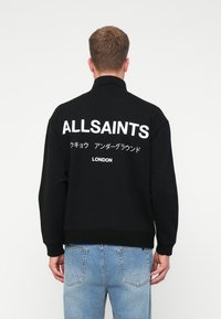 Fekete kapucnis pulóvert viselő férfi, amelyen fehér "ALLSAINTS" felirat és japán szöveg található a hátán, világoskék farmernadrággal, háttal állva.
