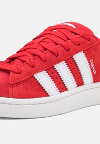 Zapatilla de ante roja con diseño de tres rayas de piel blanca, cordones texturizados, cuello acolchado y suela de goma. Texto "CAMPUS" en el lateral.