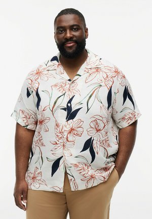 Bearded man wearing a short-sleeve white shirt with large orange floral prints and tan pants, standing with one hand in pocket.