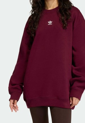 Personne portant un sweat-shirt Adidas oversize couleur bordeaux avec un logo blanc sur la poitrine, associé à un pantalon foncé, sur un fond clair uni.