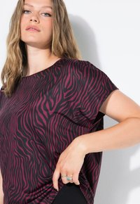 Mujer con cabello largo y rubio lleva una camiseta de manga corta a rayas en púrpura y negro, sujetando una tela cerca de la cadera, contra un fondo blanco.