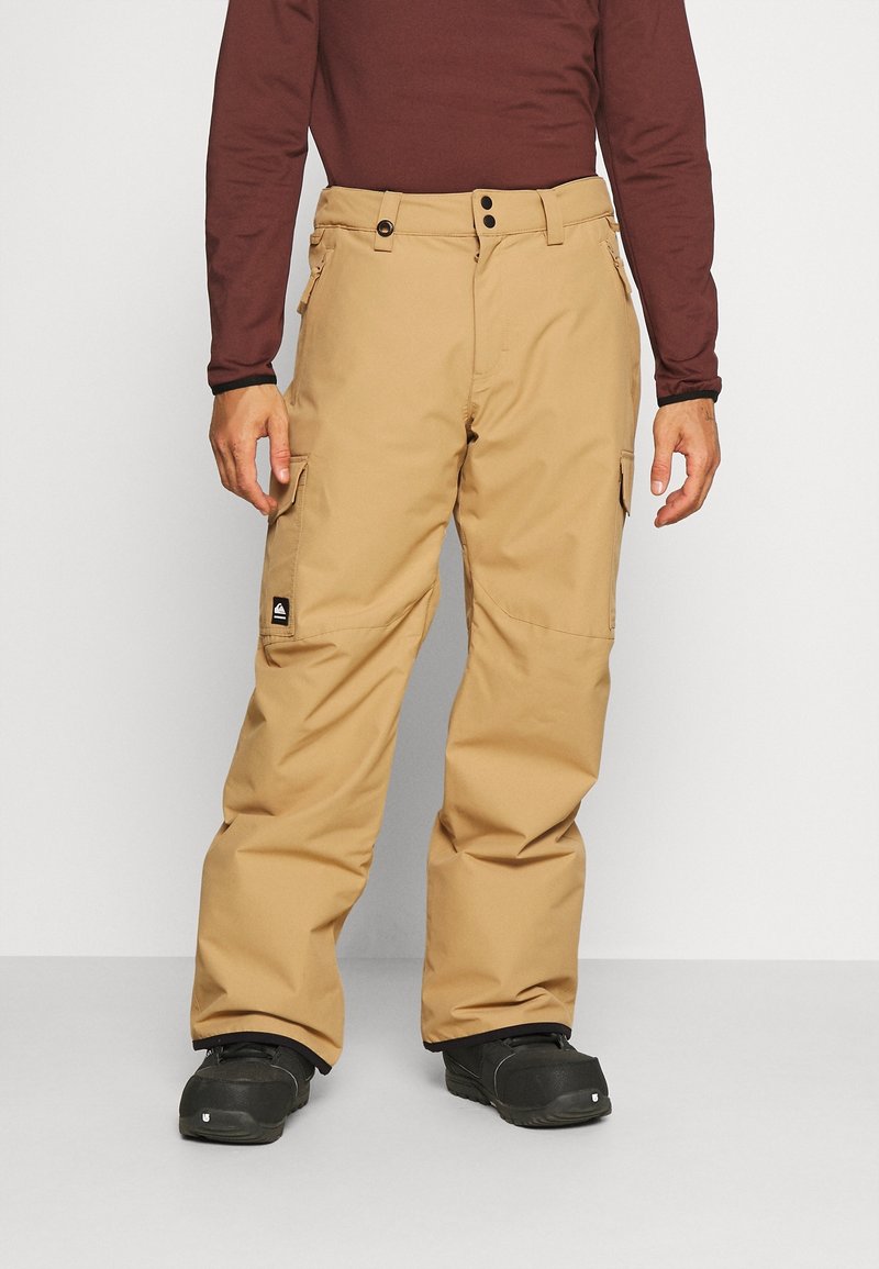 Homme portant un pantalon cargo beige, des chaussures noires et une chemise à manches longues rouge foncé, debout devant un fond neutre.