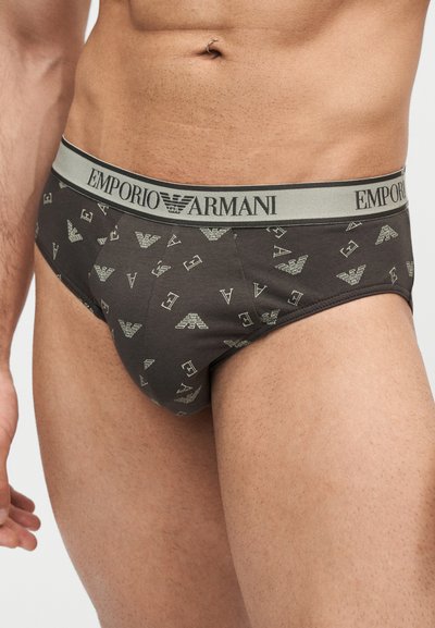Ανδρικός κορμός που φοράει σκούρα γκρι briefs Emporio Armani με ανοιχτό γκρι ζώνη μέσης και μοτίβο λογότυπου, στέκεται μπροστά σε απλό φόντο.
