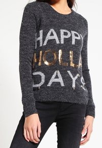 Grijze trui met de tekst "HAPPY HOLLA DAYS" in witte en gouden pailletten, met een ronde halslijn en lange mouwen, van een gebreide stof met textuur.