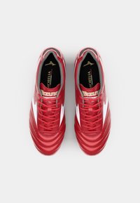 Des crampons de football rouges présentent un dessus en cuir texturé, des accents latéraux blancs et un intérieur noir. Les lacets sont rouges, avec un logo doré sur la languette.