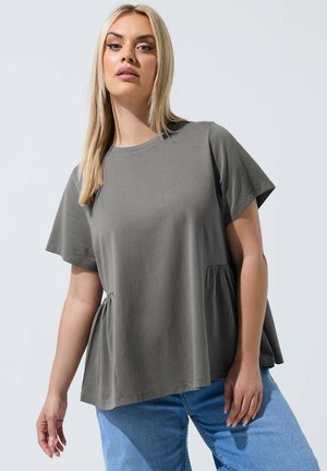 Lyshåret kvinde iført løs grå kortærmet top med sidesamlinger og blå jeans, stående mod en lysegrå baggrund.
