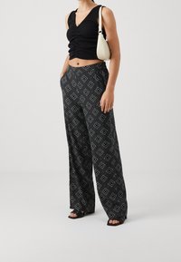 Kvinna som bär en svart rynkad crop top, mönstrade högt midjade byxor med vida ben, svarta sandaler och en liten vit axelväska.