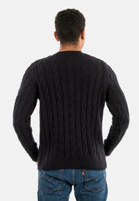 Persoon met kort krullend haar, gekleed in een zwart kabelgebreid vest en blauwe spijkerbroek, met handen op de heupen, met de rug naar de kijker gekeerd.
