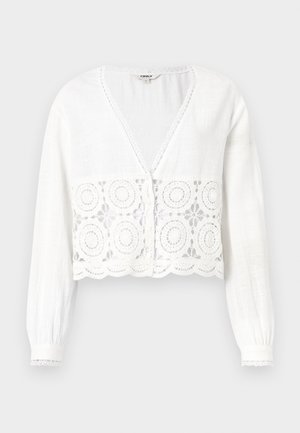 Blusa blanca corta con mangas largas, escote en V y un diseño floral de encaje en el dobladillo, con bordes de ondas y detalles de botones.