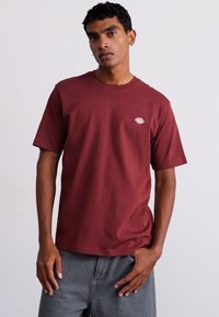 T-shirt corto color bordeaux in cotone morbido con colletto girocollo e piccolo logo ricamato nella parte superiore sinistra del petto. Pantaloni grigi.