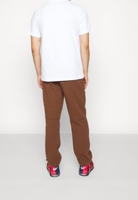 Man står med ryggen mot kameran iförd en enkel vit kortärmad skjorta, bruna byxor och röda samt blå Nike-skor på en vit bakgrund.