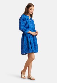 Blue dress with floral embroidery, long sleeves, gathered waist, knee-length, paired with light sandals. Textured, lightweight fabric.