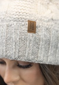 Gorro de punto en gris claro con textura acanalada y una pequeña etiqueta de cuero rectangular. El material parece suave y grueso.
