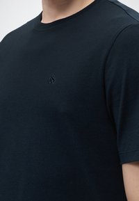 Primer plano de una persona que lleva una camiseta negra lisa con cuello redondo y un pequeño logotipo bordado en el pecho izquierdo.