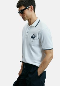 Man met zwarte zonnebril, witte polo met zwart logo en bies, en zwarte broek met handen in de zakken, tegen een effen achtergrond.