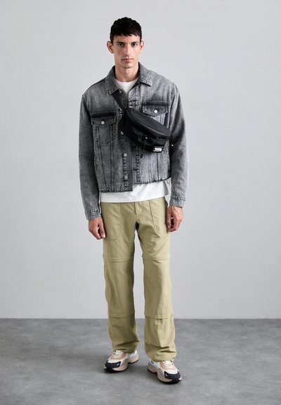 Veste en denim gris clair avec des poches poitrine doubles, un t-shirt blanc ajusté en dessous, un pantalon olive et des baskets multicolores avec des accents sombres.