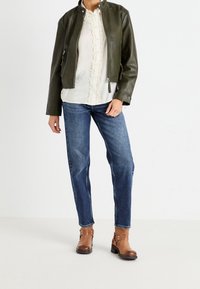 Chaqueta de cuero verde oliva con cuello alto, blusa blanca de botones con detalles de encaje, vaqueros de mezclilla azules y botas de tobillo marrones.