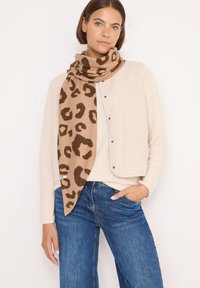 Beige fleece vest med trykknaplukninger, parret med et brunt leopardprint tørklæde. Modellen har blå denim jeans på med en afslappet pasform.