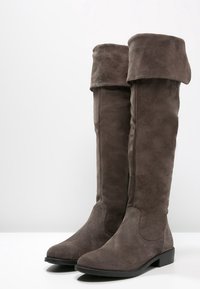 Bottes hautes en daim gris foncé avec des revers pliés et de petits talons noirs, posées côte à côte sur une surface et un fond blancs.