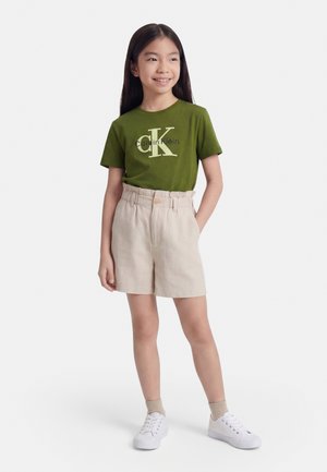 Tüdruk pikkade tumedate juustega, kandes oliivirohelist Calvin Kleini T-särki, beežisid lühikesi pükse, beežisid sokke ja valgeid vabaajajalatsid, naeratades ja seistes.