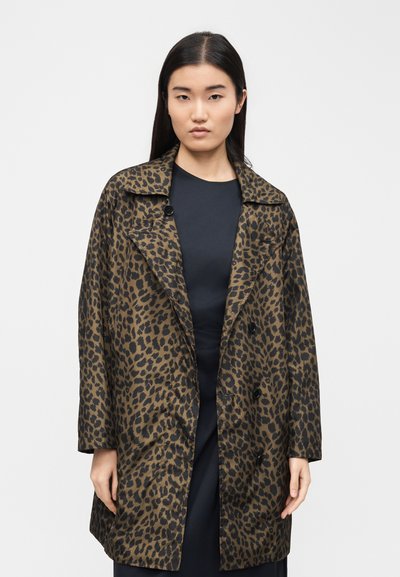 Γυναίκα με ίσια μαύρα μαλλιά, φοράει ένα καφέ και μαύρο παλτό με μοτίβο λεοπάρδαλης με κουμπιά πάνω από ένα μαύρο φόρεμα, στέκεται μπροστά σε ένα απλό φόντο.