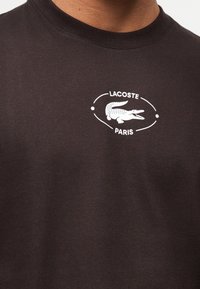 Gros plan d'une personne portant une chemise marron foncé avec un logo Lacoste Paris blanc représentant un crocodile sur la poitrine.