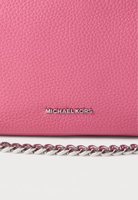 Ροζ δερμάτινη τσάντα χειρός με υφή στην επιφάνεια, με αλυσίδα από ασημί μέταλλο και ανάγλυφο λογότυπ ο με κεφαλαία γράμματα στο μπροστινό μέρος.