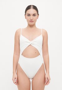 Maillot de bain une pièce blanc avec un détail de nœud à l'avant, des côtés découpés, des bretelles fines et un motif texturé sur l'ensemble du tissu.