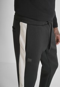 Zwarte joggers met een witte zijstreep, elastische tailleband en trekkoord. Voorzien van een klein zwart logopatch aan de voorkant. Zachte stof textuur.