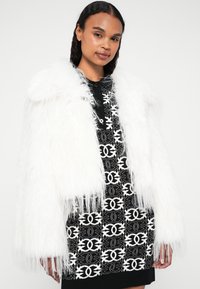 Lažna bela bunda iz umetnega krzna z dolgimi, mehkimi vlakni, nošena čez črno obleko s belim geometrijskim vzorcem in ponavljajočimi se logotipi.