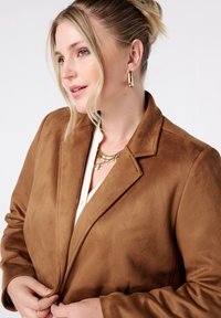 Blazer en suède marron avec des revers crantés, texture lisse et silhouette cintrée. Porté par-dessus une chemise légère à boutons, accessoirisé de bijoux en or.