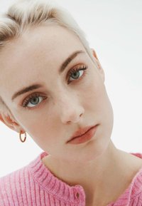 Primer plano de un rostro con piel clara, cabello rubio claro y un suéter de punto rosa. Pendiente de aro dorado, maquillaje natural y ojos azules claros.
