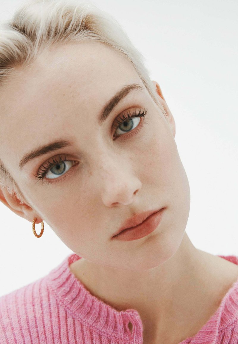 Primer plano de un rostro con piel clara, cabello rubio claro y un suéter de punto rosa. Pendiente de aro dorado, maquillaje natural y ojos azules claros.