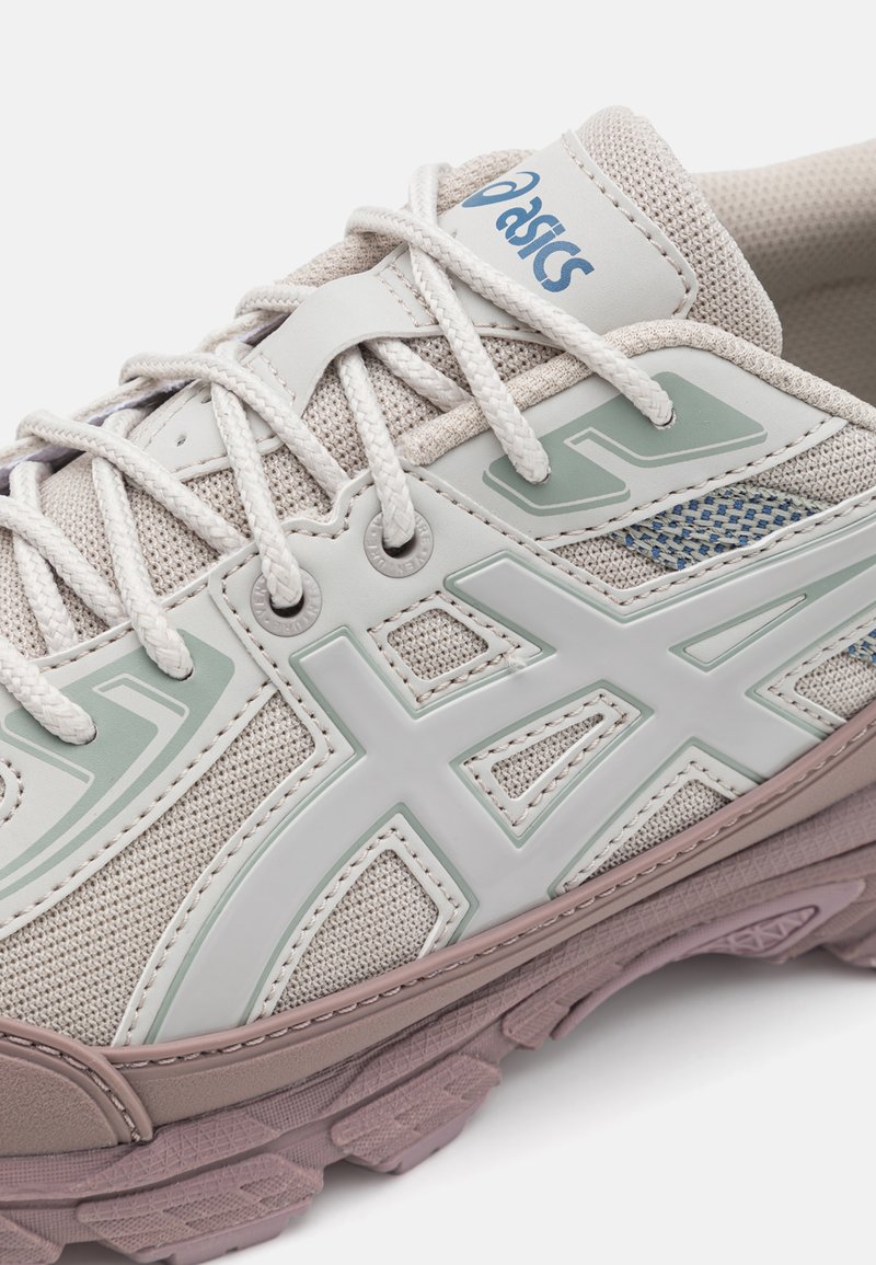 Basket de sport beige avec dessus en maille, accents vert clair et superpositions blanches. Présente une semelle en caoutchouc épaisse et des lacets dans un design robuste.