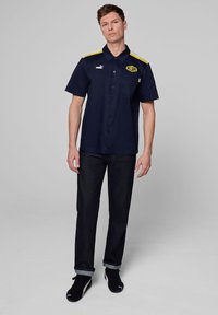 Granatowa koszula z krótkim rękawem z guzikiem z przodu, żółte akcenty na ramionach, logo Puma oraz napis "WILLIAMS" nadrukowany na klatce piersiowej; zestawiona z ciemnymi dżinsami.