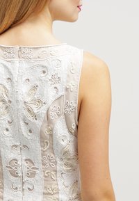 Robe sans manches beige ornée de motifs floraux et paisley en broderie blanche délicate ; le dos présente une fermeture éclair discrète.
