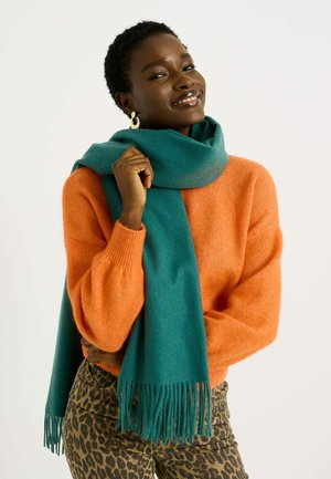 Bufanda teal con flecos drapeada alrededor del cuello, acompañada de un suéter naranja y pantalones de estampado de leopardo marrón. Textura de tela suave.