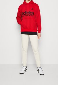 Sudadera roja con capucha y rayas negras, gran logo "adidas", combinada con pantalones joggers color crema y zapatillas en blanco y negro.