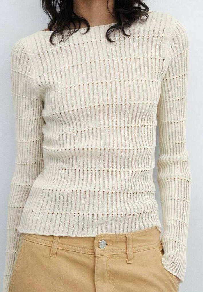 Femme portant un pull crème côtelé ajusté à manches longues, rentré dans un pantalon taille haute marron clair avec un bouton et des passants de ceinture.