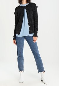 Svart, glittrig cardigan med volangärmar, knappstängning, lager på en ljusblå topp. Kombinerad med slitna blå jeans och vita stövlar.