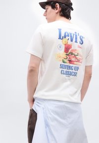 Νεαρό άτομο που φοράει λευκό T-shirt Levi’s με γραφικό και σύνθημα «Serving Up Classics» στην πλάτη, απεικονίζοντας μπέργκερ, πατάτες και ποτό.