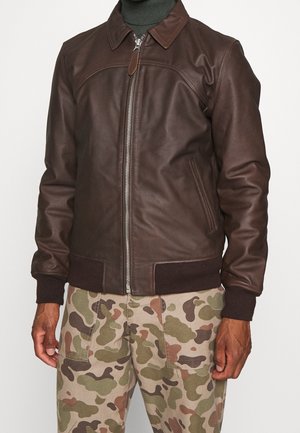 Mand iført mørkebrun læderjakke med lynlås og krave, ribstrikkede manchetter og kant, over olivengrøn rullekrave, og camouflagebukser.