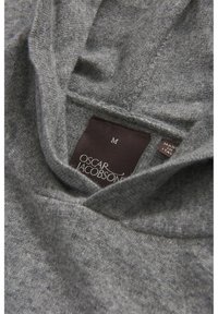 Grå sweater med blød tekstur, der har en hætte og synlige syninger. Etiketterne viser "Oscar Jacobson" og størrelse "M." Fremstillet i Italien.