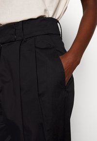 Svarta skräddarsydda byxor med veck, hög midja och sidofickor; tillverkade av ett mjukt, lätt tyg med matt yta.