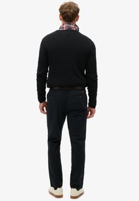 Hombre de pie de espaldas, vestido con un suéter negro, con el cuello de una camisa de cuadros visible, pantalones oscuros, cinturón marrón y zapatillas blancas con suelas de goma.