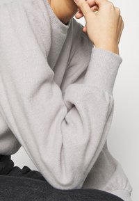 Světle šedý pletený svetr s vysokým výstřihem a objemovými rukávy. Měkká textura, volný střih a žebrované detaily na manžetách.