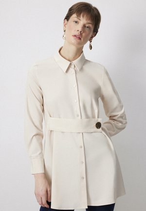 Chemise crème à boutons avec des manches longues, un col et une taille ceinturée avec un bouton d'accent doré. Tissu lisse, coupe décontractée.